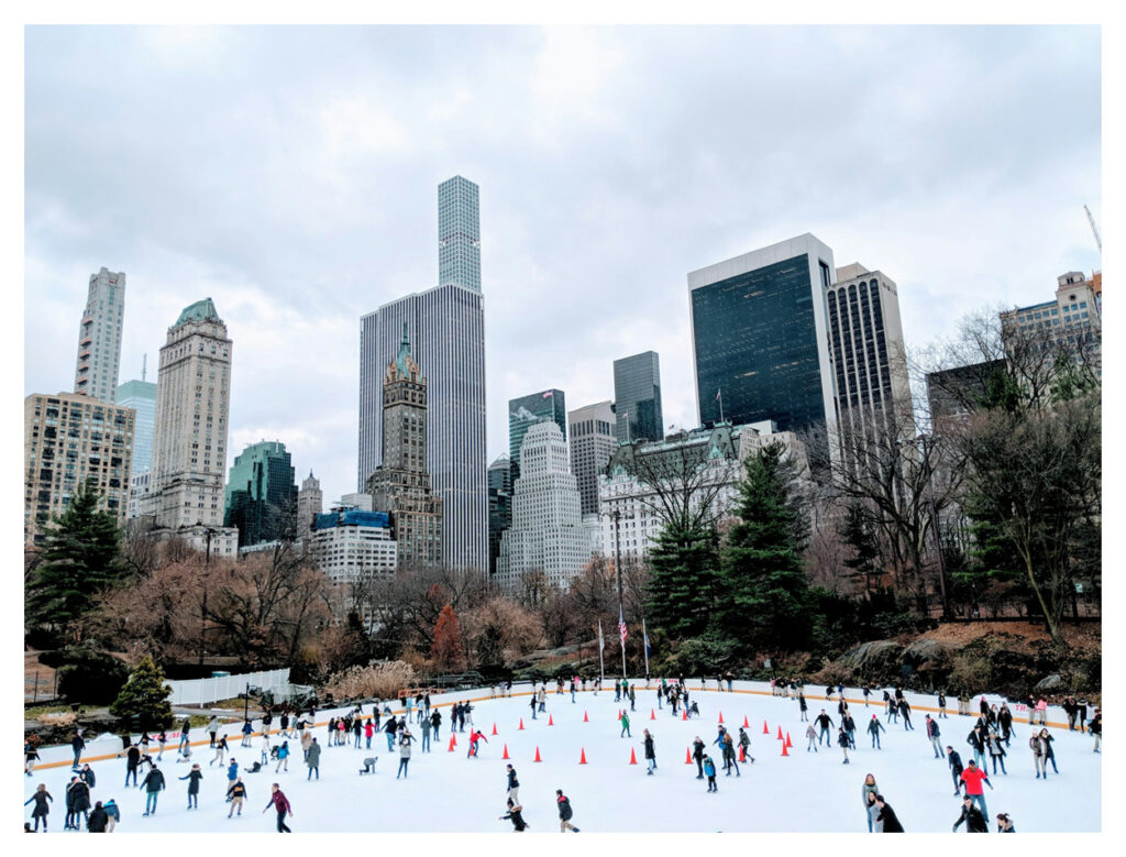 A skating rink with a bunch of people in it ice skating, there are buildings and trees around